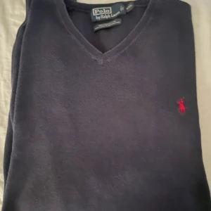 Mörkblå v-ringad tröja Polo Ralph Lauren - Snygg mörkblå v-ringad tröja från Polo Ralph Lauren i 100% pima bomull. Klassisk design med röd broderad logga på bröstet. Perfekt för dig som gillar stilrena och tidlösa plagg med premiumkänsla. Passar mer XL