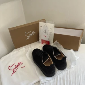 Svarta sneakers från Christian Louboutin - Säljer ett par svarta sneakers från Christian Louboutin med ikoniska röda sulor och snyggt broderad logga undertill. Skorna har snörning och är tillverkade i mocka med platt sula. Kommer med originalkartong, dustbag och äkthetsbevis.