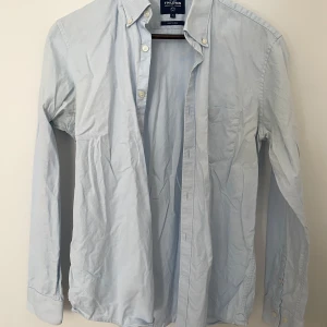 Ljusblå skjorta från T.M. Lewin - Snygg ljusblå skjorta från T.M. Lewin med klassisk krage och bröstficka. Skjortan har långa ärmar, knappar framtill och är tillverkad i mjuk bomull som är lätt att stryka. Perfekt för dig som gillar stilrena och enkla plagg.