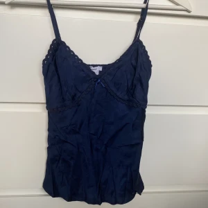 Satin linne - Supersöt mörkblå linnetopp från Subdued med tunna axelband och spetsdetaljer längs urringningen. Toppen har en liten rosett framtill och är gjord i ett glansigt material som känns lyxigt. Perfekt att styla med jeans eller kjol för en trendig look.