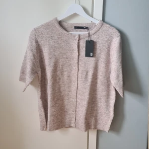 Beige kofta med pärlknappar från 157 - Supermjuk beige kofta från 157 med korta ärmar och rund hals. Koftan har snygga pärlknappar framtill och är stickad i ett fluffigt material som känns både trendigt och bekvämt. Perfekt att slänga över en topp för en chill look.