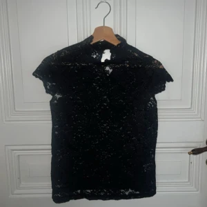 Svart spetsblus med hög krage - Säljer en svart blus i genombruten spets med hög krage och knapp i nacken. Blusen har korta ärmar och fina vågiga kanter upptill. Perfekt för dig som gillar detaljer och vill sticka ut lite extra.