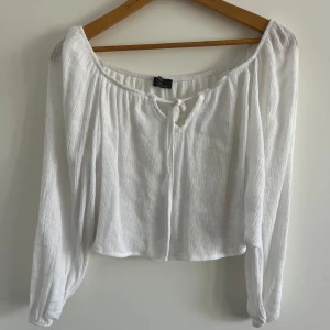 Vit croppad blus från Bershka - Superfin vit blus från Bershka med croppad passform och långa ärmar. Toppen har en offshoulder-skärning och knytning framtill vid halsen. Tunt, lätt material som ger en luftig känsla – perfekt för varma dagar eller lager på lager.