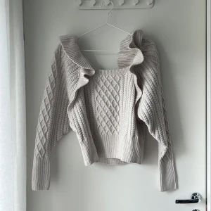 Beige stickad volangtröja H&M - Superfin beige stickad tröja från H&M med volangdetaljer längs axlarna och ett snyggt kabelstickat mönster. Tröjan har lång ärm, bred ribbad mudd och fyrkantig halsringning. Perfekt för dig som gillar en söt och trendig look.