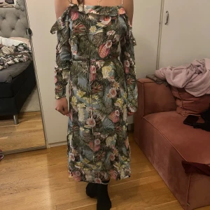 Blommig långklänning från H&M - Superfin långklänning från H&M med tropiskt blommönster i grönt, rosa, gult och blått. Klänningen har offshoulder, volangdetaljer, öppen rygg och knyt i midjan. Perfekt för sommarens alla tillfällen!