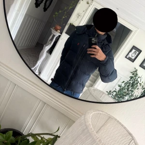 Mörkblå dunjacka Polo Ralph Lauren - Säljer en svart dunjacka från Polo Ralph Lauren med huva och dragkedja. Jackan har en klassisk pufferstil och diskret logga på bröstet. Perfekt för kalla dagar och har en snygg, enkel design som funkar till det mesta. Finns två små hål i jackan som jag har satt tejp på så inte det ska läcka men det kan man nog sy igen eller så kan man lämna in den till en skräddare annars är skicket bra nypris 6000kr och mitt pris 2000kr kvitto och påse finns om man vill ha med det så hör av er vid funderingar😊