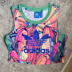 Blommig croppad linnetopp från Adidas - Färgstark croppad linnetopp från Adidas med stort blått logotryck framtill. Toppen har ett blommigt mönster i rosa, grön, orange och blå toner samt gröna kanter. Perfekt för dig som gillar sportig och unik stil.
