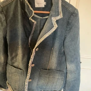 Jeanskavaj från Baravara, storlek 40 - Snygg jeanskavaj från Baravara i klassisk blå denim. Kavajen har råa, fransiga kanter och dekorativa sömmar, samt stora fickor framtill. Stängs med knappar och har en avslappnad men ändå dressad vibe.