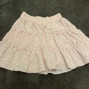 Rosa blommig volangkjol från Zara - Supersöt rosa kjol från Zara med små blommor i grönt och gult. Kjolen har flera volanger och ett smockat midjeband som gör den bekväm och lätt att bära. Perfekt för dig som gillar en feminin och lekfull stil.