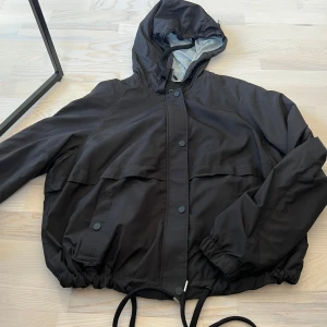 Svart vindjacka med huva från Divided - Säljer en svart vindjacka från Divided med huva och dragsnöre i nederkant. Jackan har knappar och dragkedja framtill samt två fickor med lock. Perfekt för blåsiga dagar och enkel att matcha med olika outfits.