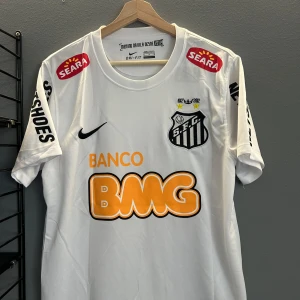 Vit Santos FC fotbollströja Neymar Jr #11 - Santos FC matchtröja i vitt med Neymar Jr och nummer 11 på ryggen. Tröjan har korta ärmar, svarta och orangea tryck samt flera sponsorer. Klassisk Nike-logga på bröstet och klubbmärke på vänster sida. En replika helt ny med tagg 