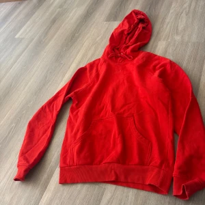 Röd hoodie med ficka - Säljer en klassisk röd hoodie med huva och magficka. Perfekt för dig som gillar en enkel och stilren look. Tröjan har långa ärmar och ribbade muddar vid ärmslut och nederkant. Passar både till skolan och chill hemma.