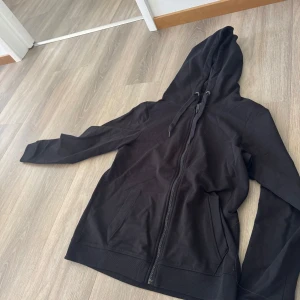 Svart hoodie med dragkedja - Svart hoodie med dragkedja och huva. Jackan har två fickor framtill och snörning i huvan. Perfekt för en avslappnad stil och enkel att matcha med det mesta.