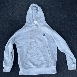 Grå hoodie från Harlaut - Säljer en grå hoodie från Harlaut apparel med ett stort blåt h tryck på ryggen och liten blå detalj på bröstet. Använd ett par fåtal gånger. Perfekt för en avslappnad stil.