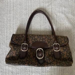 DKNY handväska  - Säljer en brun handväska från DKNY med tydligt monogrammönster och mörkbruna detaljer. Väskan har tre dekorativa spännen framtill och två rejäla handtag. Perfekt för dig som gillar klassisk och tidlös design. I bra skick 