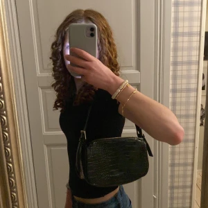 Svart axelväska i croco-mönstrat skinn från Gina Tricot - Säljer en svart axelväska från Gina Tricot i croco-mönstrat skinnimitation. Väskan har silverfärgade detaljer och dragkedja upptill. Perfekt storlek för det viktigaste och enkel att matcha till olika outfits. Den är i väldigt bra skick. Det finns en liten ficka i väskan med dragkedja som går och stänga.