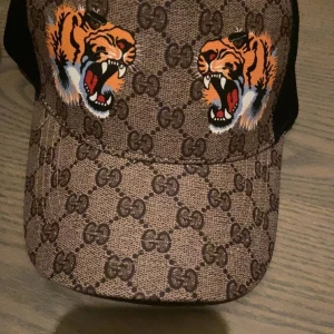 Gucci keps med tigertryck - Snygg keps från Gucci med klassiskt GG-mönster i beige och brunt samt två färgstarka tigrar framtill. Justerbar passform och svart mesh på baksidan. Perfekt för dig som vill sticka ut med en exklusiv accessoar.