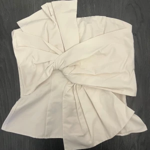 Vit axelbandslös topp med rosettdetalj - Säljer en vit axelbandslös topp i korsettstil med stor rosettdetalj framtill. Toppen har dragkedja i ryggen och är figursydd för en snygg passform. Perfekt för dig som vill sticka ut med en unik och trendig look. Lappet sitter kvar, därmed helt oanvänd.