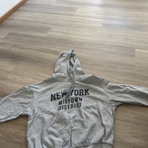 Grå hoodie med New York-tryck - Säljer en grå hoodie med trycket 'NEW YORK MIDTOWN DISTRICT' på bröstet. Tröjan har huva med dragsko och ribbade muddar vid ärmslut och nederkant. Perfekt för en avslappnad stil.