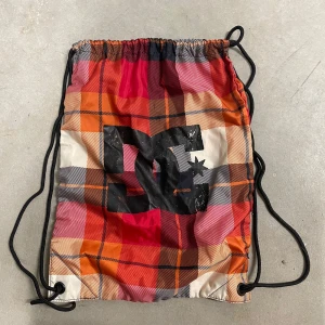 Rutig gympåse från DC - Säljer en färgglad gympåse från DC med rutigt mönster i rött, orange, svart och vitt. Väskan har dragskostängning med svarta snören och stor DC-logga på framsidan. Perfekt för träning eller vardagsäventyr.