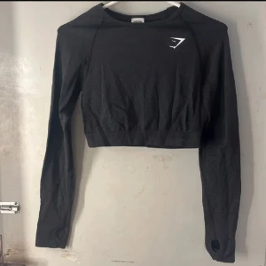  träningstopp  - Säljer en svart långärmad träningstopp från Gymshark med croppad passform. Toppen har tumhål vid ärmsluten och en diskret logga på bröstet. Perfekt för gymmet eller löpning. I detta köp ingår även sporttoppen som man kan ha under, dessa saker är använda väldigt få gånger och är super sköna i materialet. Man får båda 2 för 400 kr😊 Tröjan är i stolek M och sport bhn i S