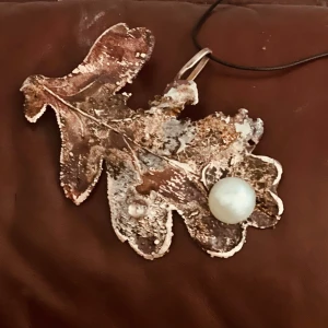 Halsband med ekblad och pärla - Unikt halsband med ett stort hänge i form av ett ekblad i metall med en stor vit pärla. Hänget har en rustik och konstnärlig känsla och hänger på ett svart band. Perfekt för dig som gillar naturinspirerade smycken.
