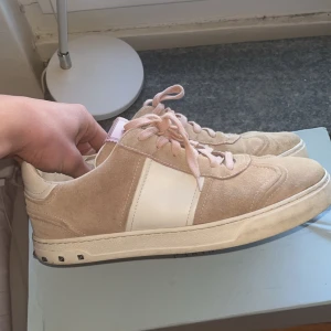Valentino Flycrew - Säljer ett par snygga Valentino Garavani sneakers i beige mocka med vita detaljer och rosa skosnören. Skorna har en vit sula med nitar baktill och en bred vit rand på sidan. Perfekta för dig som gillar exklusiva och trendiga sneakers.