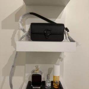 Svart axelväska från Coach - Snygg svart axelväska från Coach med ett stort C-spänne framtill. Väskan har en stilren design med lock och justerbar axelrem. Perfekt för att bära det viktigaste med stil.