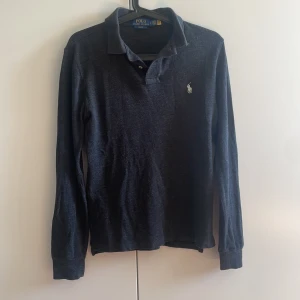 Mörkgrå långärmad pikétröja från Polo Ralph Lauren - Säljer en mörkblå långärmad pikétröja från Polo Ralph Lauren i slim fit-modell. Tröjan har klassisk krage, knappar vid halsen och den ikoniska loggan broderad på bröstet. Perfekt för en stilren och avslappnad look.