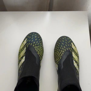 Adidas Predator fotbollsskor i svart och neon - Säljer ett par Adidas Predator fotbollsskor i svart med gula och vita detaljer. Skorna har en strumpliknande passform utan snörning och är designade för optimal bollkontroll. Perfekta för dig som vill sticka ut på planen!. Pris kan diskuteras vid snabb affär