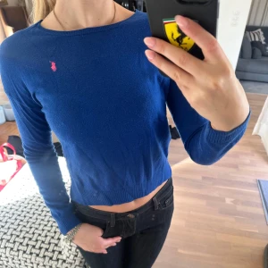 Blå stickad tröja från Ralph Lauren - Säljer en blå stickad tröja från Ralph Lauren med rund halsringning och lång ärm. Tröjan har en liten rosa logga broderad på bröstet och ribbade muddar vid ärmslut och nederkant.