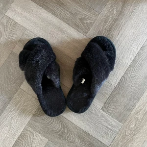 Svarta fluffiga tofflor - Mysiga svarta tofflor med korslagda band i mjukt, fluffigt material. Storlek 38-39. Knappt använda