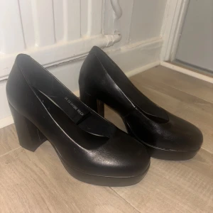 Svarta klackskor med rund tå - Säljer ett par svarta klackskor med rund tå och plattformsula. Skorna har en hög, bred klack och är tillverkade i syntetmaterial med en stilren design. Perfekta för dig som vill ha en klassisk och enkel look.