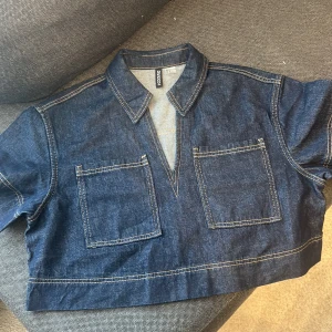 Mörkblå croppad jeansskjorta från H&M DIVIDED - Säljer en mörkblå croppad jeansskjorta från H&M DIVIDED. Skjortan har två stora bröstfickor, klassisk krage och synliga kontrastsömmar. Perfekt för vår och sommar. Skjortan är använd 1 gång 
