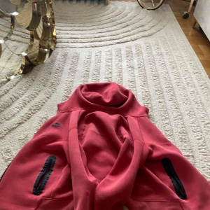 Röd nike tech fleece  - Säljer en röd hoodie från Nike med svarta dragkedjor på fickorna och svart Nike-logga på bröstet. Jackan har huva och är tillverkad i mjukt bomullsmaterial. Perfekt för dig som gillar sportig stil.
