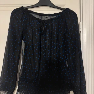 Svart blus med blå prickar och knytband - Säljer en svart transparent blus med blå prickar och knytband vid halsen. Blusen har långa ärmar och resår vid ärmslut och nederkant för en luftig och söt look. Lite genomskinlig men snygg. Använd 1 gång! Går att ha på sig den som en off-shoulder blus. 