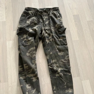 Cargobyxor i camouflage  - Säljer ett par cargobyxor i camouflage. Byxorna har elastisk midja med dragsko och stora fickor på sidorna. Perfekta för en avslappnad och cool stil.