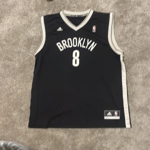Brooklyn Nets baskettröja #8 Williams - Säljer en svart Brooklyn Nets baskettröja från Adidas med nummer 8 och namnet Williams på ryggen. Tröjan har vita detaljer och är ärmlös med klassisk basketdesign. Perfekt för basketfans som vill sticka ut på planen eller på läktaren.