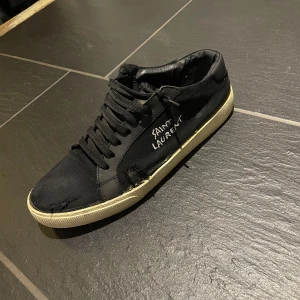 Svarta sneakers från Saint Laurent - Svarta sneakers från Saint Laurent med vit sula och broderad logga på sidan. Skorna har snörning och är tillverkade i textil och skinn. Klassisk rund tå och platt sula. Perfekta för dig som gillar stilrena och ikoniska märken.                                Har varit hos skomakaren för cirka en vecka sedan(se bild 4) där av priset till skillnad från andra på Plick.                                                                 Har tyvär inte orginal lådan kvar 