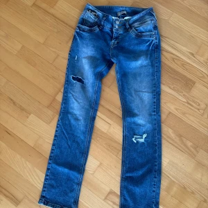 Blå raka jeans med slitningar - Säljer ett par blå jeans med raka ben och slitna detaljer på låret och knät. Jeansen har klassisk femficksdesign och normal passform. Perfekta för en avslappnad och trendig look.