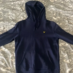 Mörkblå hoodie från Lyle & Scott - Säljer en stilren mörkblå hoodie från Lyle & Scott med klassisk huva och magficka. Tröjan har ett broderat gult logomärke på bröstet och är tillverkad i mjukt material. Perfekt för en avslappnad look.