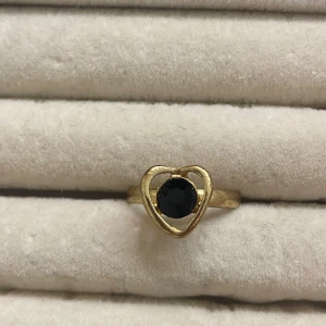 Guldfärgad ring med hjärtdesign och svart sten - Snygg guldfärgad ring med ett hjärtformat motiv och en svart rund sten i mitten. Ringen har en enkel och stilren design som passar perfekt för dig som gillar unika smycken. Den är justerbar💞 priset går att diskutera!