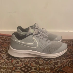 Ljusgrå Nike sneakers med vit sula - Säljer ett par ljusgrå Nike sneakers med vit sula och svart detalj vid hälen. Skorna har klassisk snörning och stor vit swoosh på sidan. Perfekta för träning eller vardagsstil.