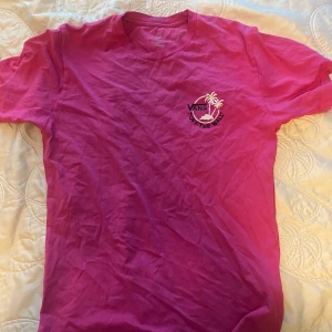Rosa t-shirt från Vans med palmtryck - Säljer en rosa t-shirt från Vans i 100% bomull. T-shirten har ett tryck med palmer och texten 'Vans Off The Wall' både fram och bak. Klassisk passform och rund halsringning. Perfekt för en avslappnad stil.