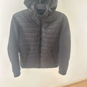 Svart quiltad jacka med huva från Lindbergh - Snygg svart quiltad jacka från Lindbergh med huva och dragkedja framtill. Jackan har två fickor med dragkedja och en diskret logga på ärmen. Perfekt nu för våren. Har ni några frågor är de bara och skriva. Pris går och diskutera.