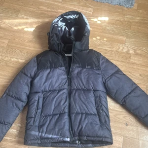 Svart och mörkgrå pufferjacka med huva - Säljer en svart och mörkgrå pufferjacka från Everest med huva och dragkedja. Jackan har två fickor med dragkedja och quiltad design. Perfekt för kyliga dagar och har en sportig look.jävligt snygg.perfekt.den kommer hålla jätte länge. Den är för höst och vinter och när det blåser lite.