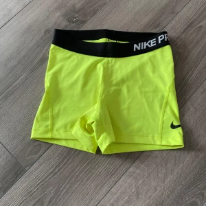 Nike Pro shorts - Säljer ett par neongula träningstights shorts från Nike Pro med svart resår i midjan och Nike-logga på benet. Perfekta för intensiva träningspass eller sportaktiviteter. Sitter tight och har en sportig look.