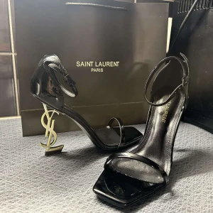 Svarta Saint Laurent klackar med YSL-detalj - Säljer ett par svarta Saint Laurent klackar i lack med öppen tå och ikonisk YSL-logga som klack. Snygg minimalistisk design med smal rem över tårna och runt vristen. Perfekta för dig som vill sticka ut med lyxig känsla.