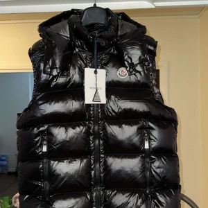 Svart dunväst från Moncler - Snygg svart dunväst från Moncler med glansig finish och huva. Västen har dragkedja framtill, två fickor med dragkedja och Moncler-logga på bröstet. Perfekt för lager-på-lager under kyliga dagar.