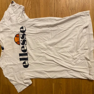 Vit t-shirt från Ellesse - Vit t-shirt från Ellesse med stor logga i mörkblått och orange framtill. Klassisk passform och rund halsringning. Perfekt för en sportig och avslappnad stil.